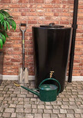 City Just Water 210L Water Butt - Black Slimline Garden Tank next to watering can and shovel on cobblestone floor