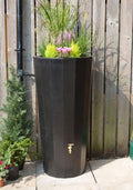 Black Metropolitan 255L Water Butt Planter - Decorative Tank with plants and spigot against a wooden fence