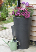 Millstone City Water Butt Planter - 180L Stone Effect Tank with blooming flowers and a green watering can.