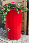 Red Midi Metropolitan Water Butt Planter - 165L Red with flowers and greenery on top, placed against a wall.
