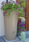 Metropolitan 255L Water Butt Planter - Sandstone Effect with blooming flowers and a watering can beside it.